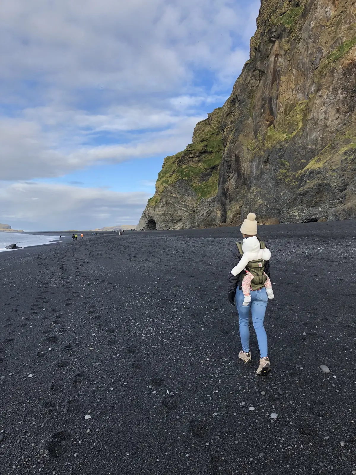 Kailah carrying our baby in a front carrier on a black sand beach below sea cliffs on Iceland's south coast
