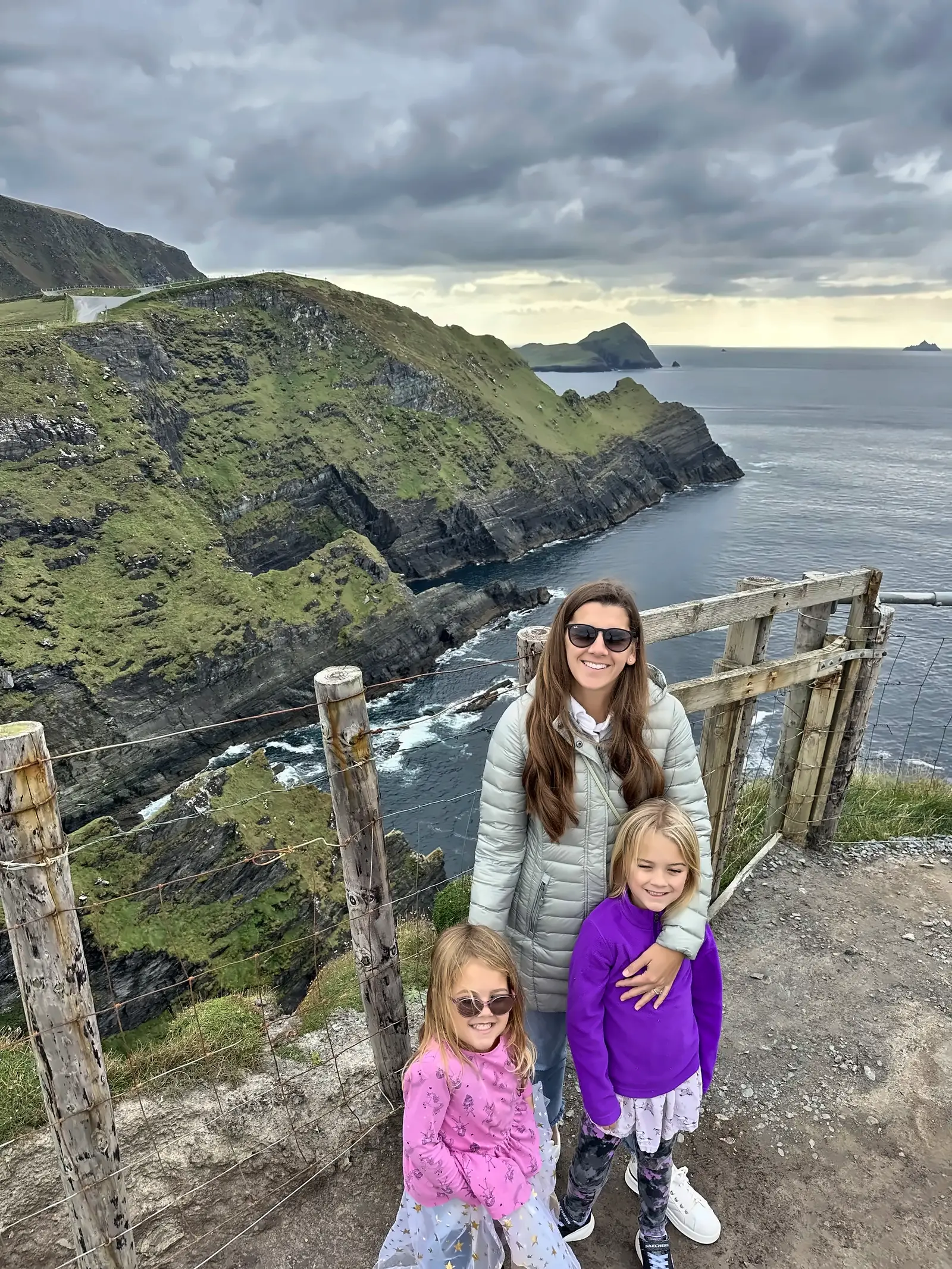 Kailah and our two daughters at a fence on the cliffs of Ireland's west coast, with sea cliffs and the Atlantic behind