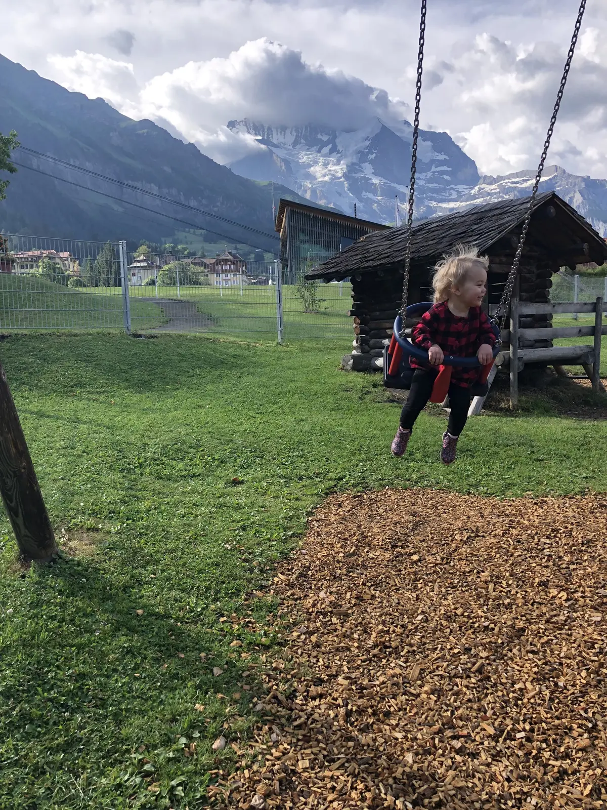 Our toddler on a playground swing with snowy Swiss mountains behind