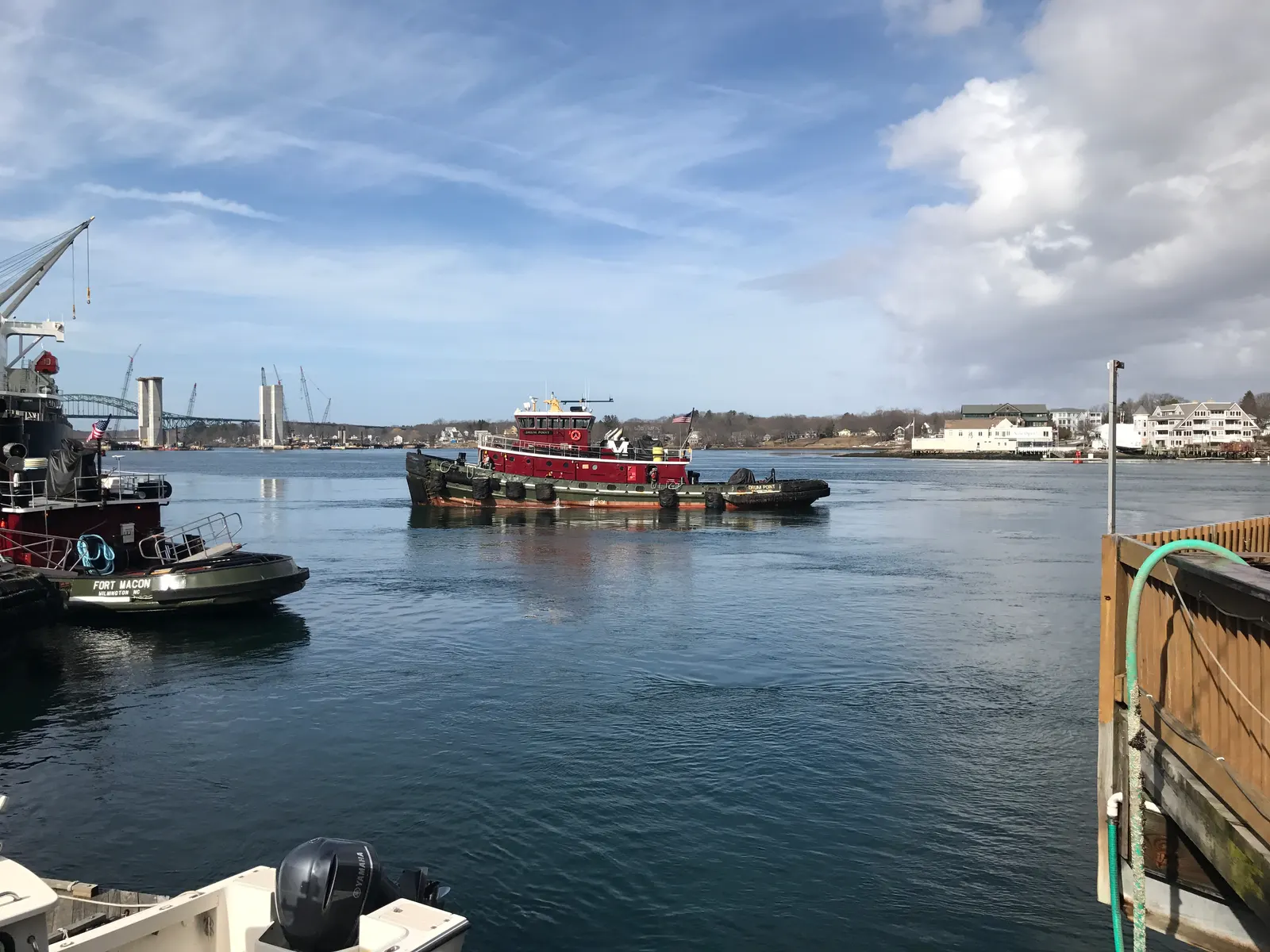 A red tugboat crossing the Piscataqua River off the Portsmouth waterfront, with docked tugs in the foreground