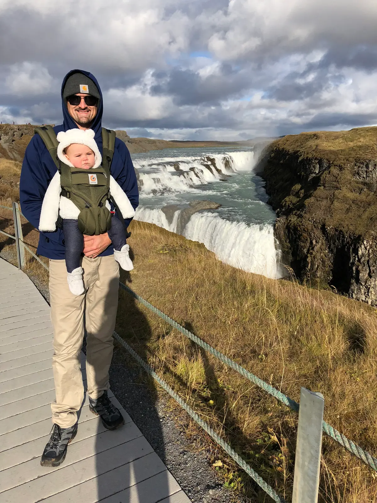 Kyle carrying our baby in a front carrier on the boardwalk above Gullfoss, the falls and brown autumn grass behind them